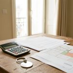 Comment calculer la taxe foncière d’un investissement locatif ? Intérieur lumineux d’un appartement locatif français avec une calculatrice, des documents administratifs vierges et des plans cadastraux simplifiés sur une table en bois.