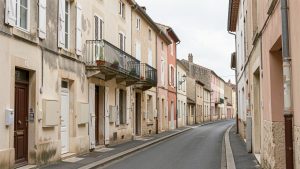 Rue résidentielle du centre-ville de Tonneins avec façades anciennes, volets, balcons et panneaux immobiliers vierges, éclairée par une lumière naturelle douce.